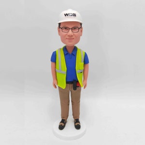 May include: A custom bobblehead figurine of a person wearing a white hard hat with the logo 'WDS' on it, a neon green safety vest over a blue collared shirt, khaki pants, and dark shoes. The figurine is standing on a white base.