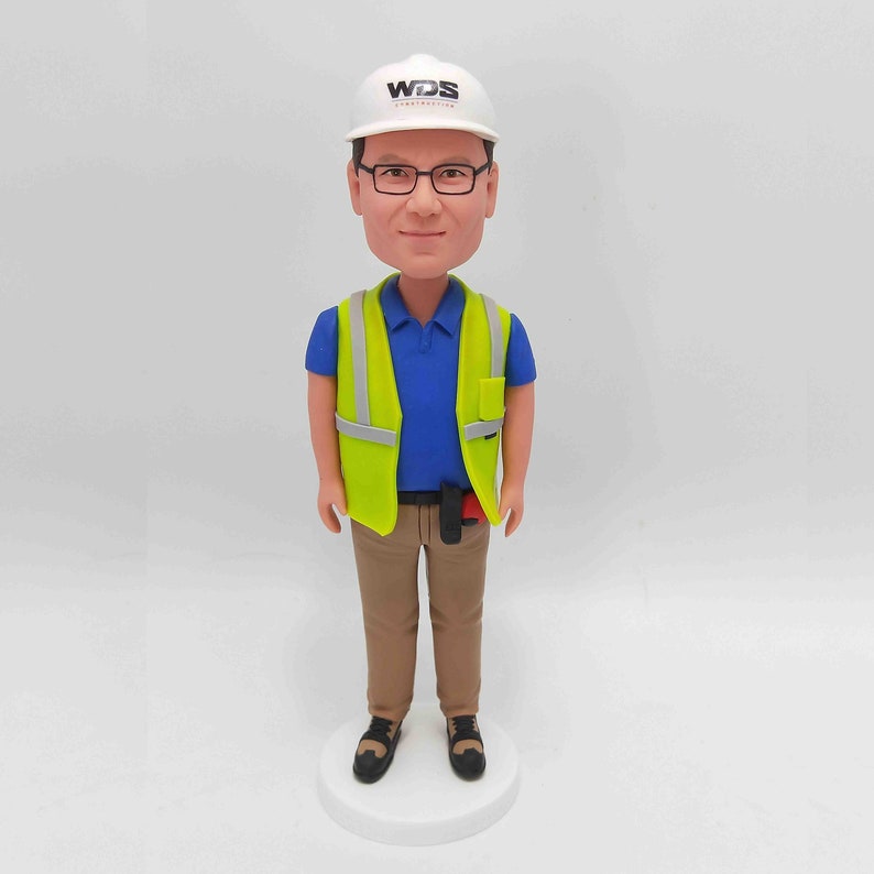 May include: A custom bobblehead figurine of a person wearing a white hard hat with the logo 'WDS' on it, a neon green safety vest over a blue collared shirt, khaki pants, and dark shoes. The figurine is standing on a white base.