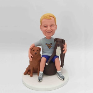 May include: A custom bobblehead figurine of a person with blond hair wearing a gray shirt with a West Virginia University logo and blue shorts, seated with two dogs. One dog is brown and the other is dark brown and white. The figurine is on a white circular base.