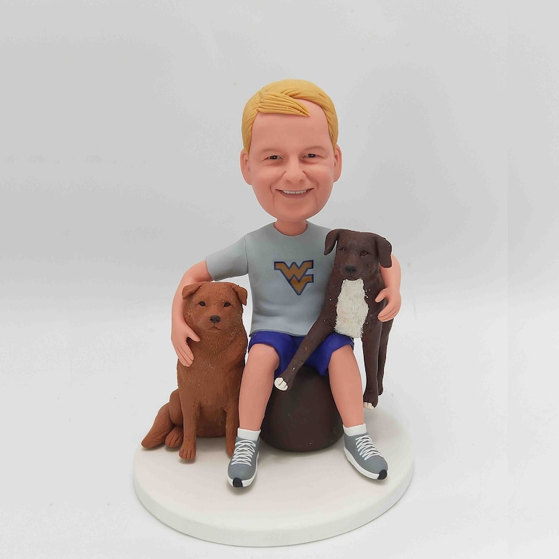 May include: A custom bobblehead figurine of a person with blond hair wearing a gray shirt with a West Virginia University logo and blue shorts, seated with two dogs. One dog is brown and the other is dark brown and white. The figurine is on a white circular base.