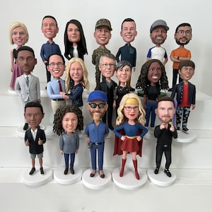 May include: A collection of personalized bobblehead figurines, each depicting a person with unique clothing and hairstyles. The figurines are arranged on tiered white platforms, showcasing a variety of poses and expressions.