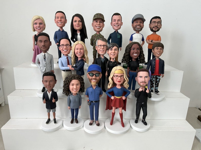 May include: A collection of personalized bobblehead figurines, each depicting a person with unique clothing and hairstyles. The figurines are arranged on tiered white platforms, showcasing a variety of poses and expressions.