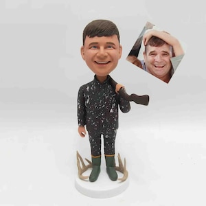 May include: A custom bobblehead figurine of a person in a camouflage jumpsuit, holding a rifle. The figurine has a smiling face and is standing on a white base with antlers. A small photo is included.