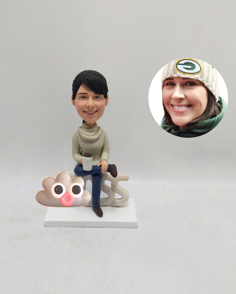 May include: A custom bobblehead figurine of a person sitting on a white base with the word "BOSS". The figurine is holding a coffee mug. A small cloud with eyes and a pink mouth is on the base. A photo of the person is in the background.