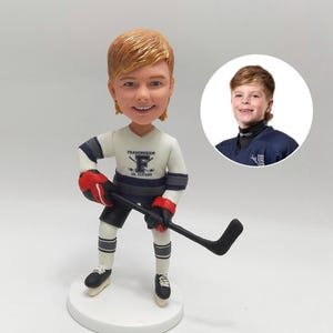 May include: A bobblehead figurine of a smiling person in a hockey uniform, holding a hockey stick. The uniform is white with blue and gray accents and the text "Framingham Jr. Flyers". A smaller image of the person is in the background.