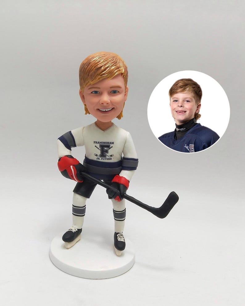 May include: A bobblehead figurine of a smiling person in a hockey uniform, holding a hockey stick. The uniform is white with blue and gray accents and the text "Framingham Jr. Flyers". A smaller image of the person is in the background.