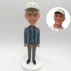 May include: A bobblehead figurine of a man wearing a light blue jacket, patterned shirt, gray pants, and black shoes. He wears a cap with text and glasses. A smaller image of the man in a cap is to the right.
