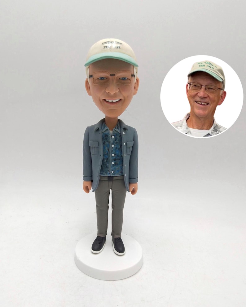 May include: A bobblehead figurine of a man wearing a light blue jacket, patterned shirt, gray pants, and black shoes. He wears a cap with text and glasses. A smaller image of the man in a cap is to the right.