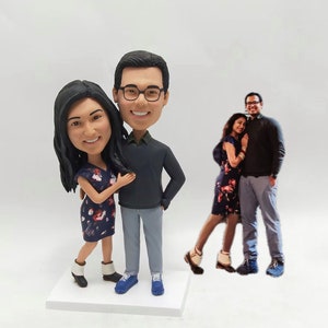 May include: Custom bobblehead figurines of a couple. The male figurine wears a dark gray sweater, gray pants, and blue shoes. The female figurine wears a dark blue floral dress and white ankle boots.  Both figurines have dark hair. A photograph of the couple is shown in the background. The bobbleheads are displayed on a white base.