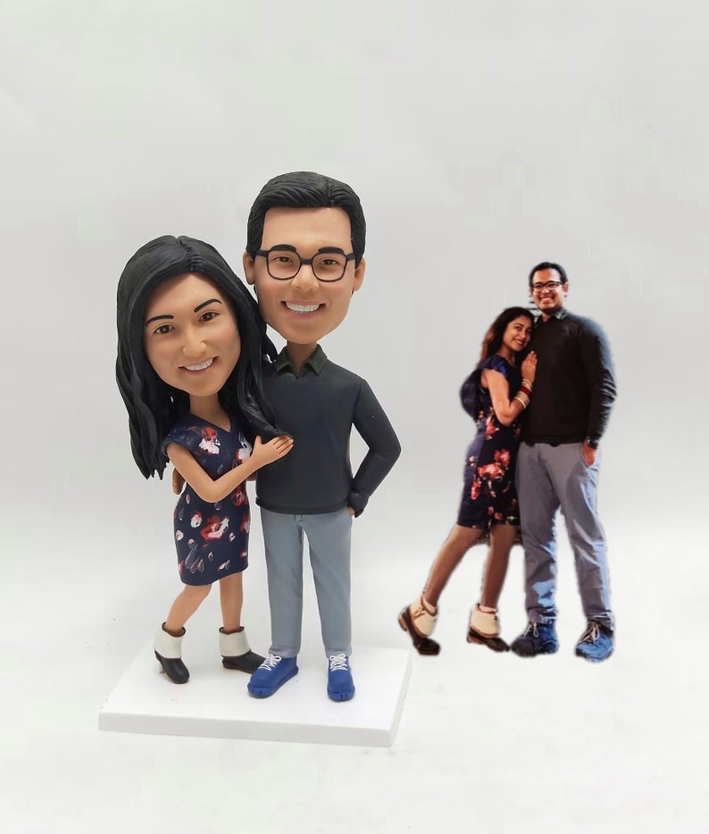 May include: Custom bobblehead figurines of a couple. The male figurine wears a dark gray sweater, gray pants, and blue shoes. The female figurine wears a dark blue floral dress and white ankle boots.  Both figurines have dark hair. A photograph of the couple is shown in the background. The bobbleheads are displayed on a white base.