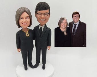May include: A custom-made cake topper featuring a couple in formal attire. The man is wearing a black suit with a white shirt and a black tie. The woman is wearing a black dress with a gold necklace. The couple is standing on a white cake base.