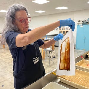 May include: A person wearing blue gloves holds a white sheet of paper with a brown image on it. The image is a photograph of a building. The person is standing in a room with a blue cabinet and a stainless steel counter. The person is wearing a black apron with the text "1 do SUN PHOTOGRAPHY" and a fern logo.