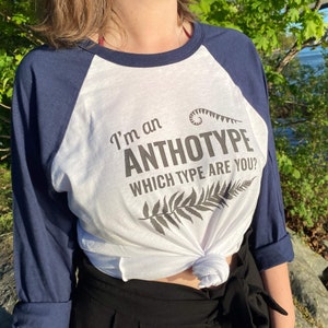 May include: A white and navy blue baseball tee shirt with the text "I'm an Anthotype Which Type Are You?" printed on the front. The shirt features a fern design.