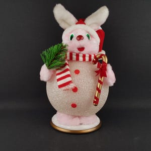 May include: A pink and white Christmas bunny figurine with a red and white striped scarf, a red hat, and a candy cane. The bunny holds a small green sprig and has red button accents on its body. The figurine is on a gold-trimmed base.