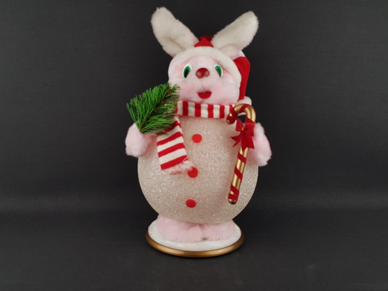 May include: A pink and white Christmas bunny figurine with a red and white striped scarf, a red hat, and a candy cane. The bunny holds a small green sprig and has red button accents on its body. The figurine is on a gold-trimmed base.