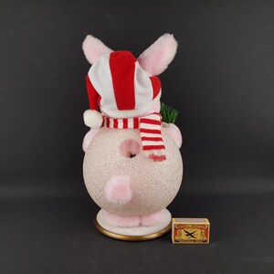 May include: A pink and white Christmas pig ornament with a red and white striped hat and scarf. The pig has a glittery body and is holding a small green sprig. A matchbox is next to the ornament.