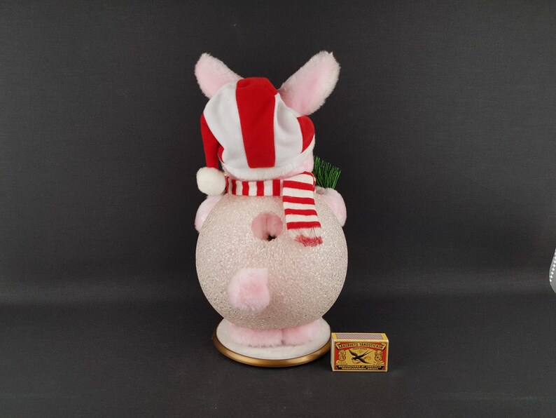 May include: A pink and white Christmas pig ornament with a red and white striped hat and scarf. The pig has a glittery body and is holding a small green sprig. A matchbox is next to the ornament.