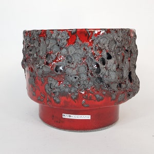 May include: Red ceramic planter with a rough, textured surface and black speckles. The planter has a wide, round base and a slightly flared top. The bottom of the planter is marked with the text "Otto Keramik".