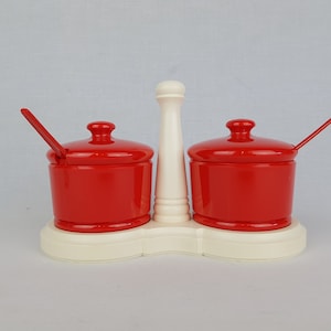 Peut inclure: Un porte-épices en céramique blanche avec deux pots en céramique rouge et des cuillères. Le support a une section centrale surélevée avec un pilier en céramique blanche.