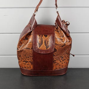Peut inclure: Sac à bandoulière en cuir marron avec un motif peau de serpent. Le sac a une fermeture à rabat et un haut à cordon. La bandoulière est en cuir marron et fixée au sac avec des anneaux argentés. Le sac mesure environ 30 cm de haut et 25 cm de large.