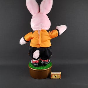 May include: A pink plush rabbit doll wearing an orange jacket and black shorts, standing on a brown pot with green faux grass. The doll has red and white shoes. A matchbox is next to the pot.