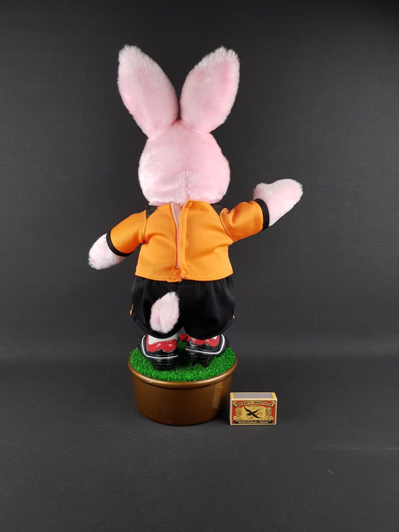 May include: A pink plush rabbit doll wearing an orange jacket and black shorts, standing on a brown pot with green faux grass. The doll has red and white shoes. A matchbox is next to the pot.