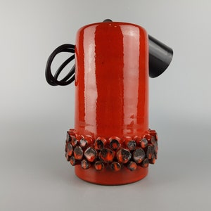 Könnte beinhalten: Eine rote Keramik-Tischlampe mit schwarzem Kabel und schwarzem Schirm. Der Sockel der Lampe hat ein dekoratives Band aus schwarzer und roter Keramik.