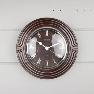 May include: A round wall clock with a dark brown frame featuring concentric circular ridges. The clock face is dark brown with white Roman numerals, white hour and minute hands, and the text Junghans, Quartz, and Made in Germany printed on it.