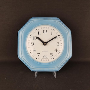 Puede incluir: Un reloj de pared octogonal con un marco azul claro que presenta un diseño de borde acanalado. La esfera es blanca con números arábigos negros, manecillas negras y el texto QUARZ y MADE IN GERMANY impreso en el centro de la esfera.