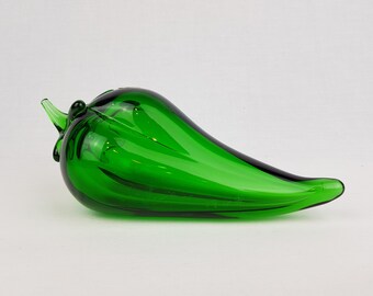 May include: A vibrant emerald green glass sculpture shaped like a chili pepper. The object features a smooth, translucent surface with a glossy finish, a curved body, and a small, pointed stem detail at the top, reflecting light across its rounded form.