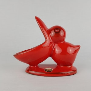 Puede incluir: Figura de pelícano de cerámica roja brillante con el pico abierto, sobre una base redondeada. El cuerpo del ave tiene curvas suaves y un ojo pequeño y detallado. Es un objeto decorativo.