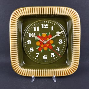 Peut inclure: Une horloge murale carrée avec un cadre en céramique vert olive brillant et une bordure beige côtelée. Le cadran est vert olive avec des chiffres arabes blancs, un motif floral central orange et vert, et l'inscription "QUARTZ" et "MADE IN GERMANY".