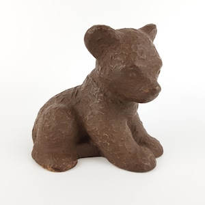 Könnte beinhalten: Eine kleine, massive braune Figur eines sitzenden Bärenjungen. Die Skulptur weist eine texturierte, unebene Oberfläche am gesamten Körper auf, mit abgerundeten Ohren, einer ausgeprägten Schnauze und einer Haltung, bei der der Bär aufrecht sitzt.