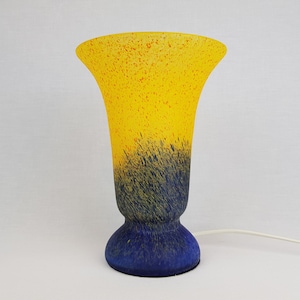 Puede incluir: Una lámpara de mesa de vidrio amarilla y azul con un diseño moteado. La lámpara tiene una base ancha y acampanada y un cuello estrecho y cilíndrico.
