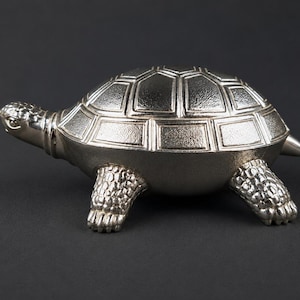 Könnte beinhalten: Eine silberfarbene Schildkrötenfigur mit detailliertem Panzer, Kopf und Beinen. Die Figur besteht aus glänzendem Metall und wird auf dunklem Hintergrund präsentiert. Der Schildkrötenpanzer hat ein geometrisches Muster.