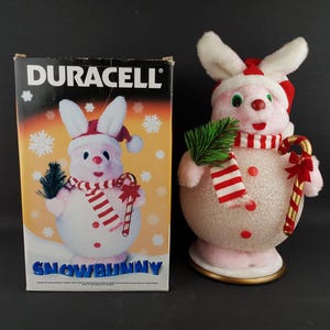 May include: A pink and white Snowbunny Christmas decoration with a red and white striped scarf, a candy cane, and a sprig of green. The box next to it has the Duracell logo and a picture of the Snowbunny.