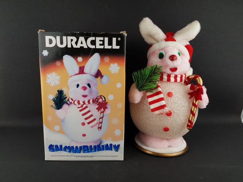 May include: A pink and white Snowbunny Christmas decoration with a red and white striped scarf, a candy cane, and a sprig of green. The box next to it has the Duracell logo and a picture of the Snowbunny.