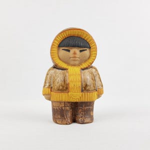 Könnte beinhalten: Eine Keramikfigur einer Person in einem Kapuzenparka. Der Parka ist beige mit einer gelben Kapuze und Besatz. Die Figur hat schwarzes Haar und eine braune Hose. Die Figur steht aufrecht.