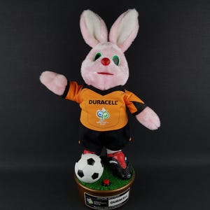 May include: A pink plush rabbit wearing an orange and black soccer uniform with the Duracell logo. The rabbit is standing on a base with a soccer ball and a red button. The base has text that reads "Duracell".