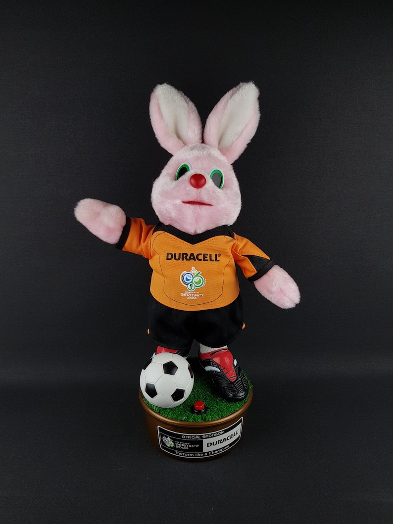 May include: A pink plush rabbit wearing an orange and black soccer uniform with the Duracell logo. The rabbit is standing on a base with a soccer ball and a red button. The base has text that reads "Duracell".