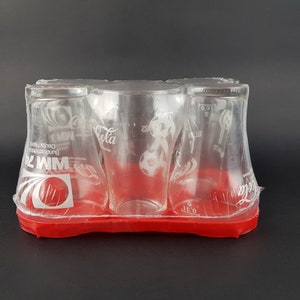 Puede incluir: Conjunto de cuatro vasos de vidrio transparente Coca-Cola con un soporte de plástico rojo. Los vasos presentan un logotipo de Coca-Cola blanco y un diseño de balón de fútbol. El soporte tiene el texto "WM 74" y "Bundesrepublik Deutschland" impreso.