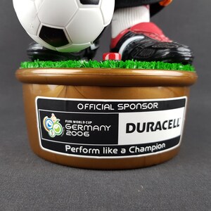 May include: A figurine of a soccer player with a soccer ball, wearing black and red cleats. The base is brown with a sticker that says "OFFICIAL SPONSOR" and "DURACELL" with the FIFA World Cup Germany 2006 logo. The text "Perform like a Champion" is also visible.