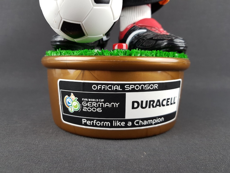 May include: A figurine of a soccer player with a soccer ball, wearing black and red cleats. The base is brown with a sticker that says "OFFICIAL SPONSOR" and "DURACELL" with the FIFA World Cup Germany 2006 logo. The text "Perform like a Champion" is also visible.
