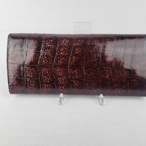 May include: A brown, rectangular wallet with a crocodile skin texture. The wallet is a dark shade of brown with a glossy finish. The wallet is displayed on a white surface.