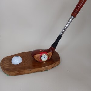 May include: A wooden golf club ornament with a clock face and a golf ball on a wooden base. The club has a brown head and handle with a silver shaft. The golf ball is white with the word "SRIXON" printed on it.