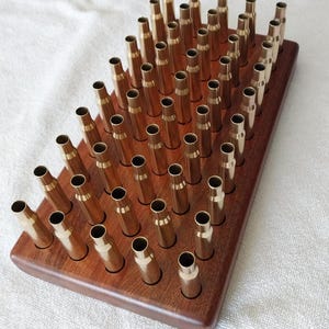 May include: A rectangular dark brown wooden block featuring a grid of circular holes, each holding a metallic brass cartridge casing. The brass casings are arranged in multiple rows and columns, standing upright in the wooden base.
