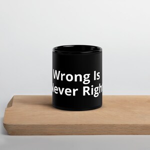 Peut inclure: Une tasse en céramique noire avec l'inscription blanche "Wrong Is Never Right". La tasse cylindrique est posée sur une planche en bois brun clair.
