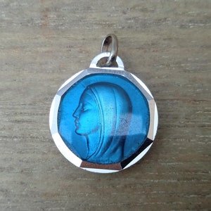 Puede incluir: Un colgante de plata con un inserto de vidrio azul que presenta un perfil de la Virgen María.