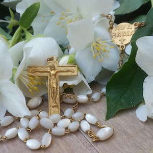 Puede incluir: Un rosario de oro con cuentas blancas y un crucifijo, descansando sobre una superficie de madera con flores blancas de jazmín.