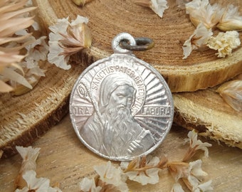 May include: Silver pendant with a depiction of a saint, surrounded by a sunburst design. The inscription reads "Sanctus Pater Benedictus Ora et Labora".